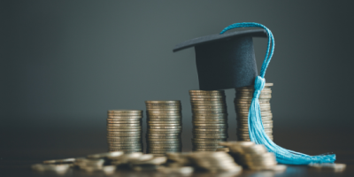 A tiny graduation cap with a blue tassle sits on a pile of quarters